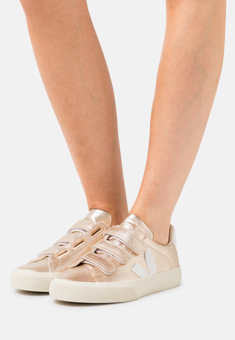 Metalne rose gold tenisice s čičak trakama, bijelim logotipom i teksturiranom gumom, s niskim dizajnom.
