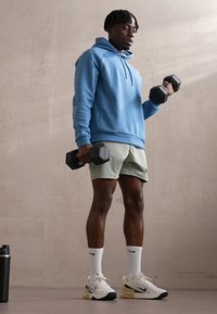 Man in blue hoodie and shorts holding dumbbells, standing on gray floor against beige wall, with black water bottle nearby.