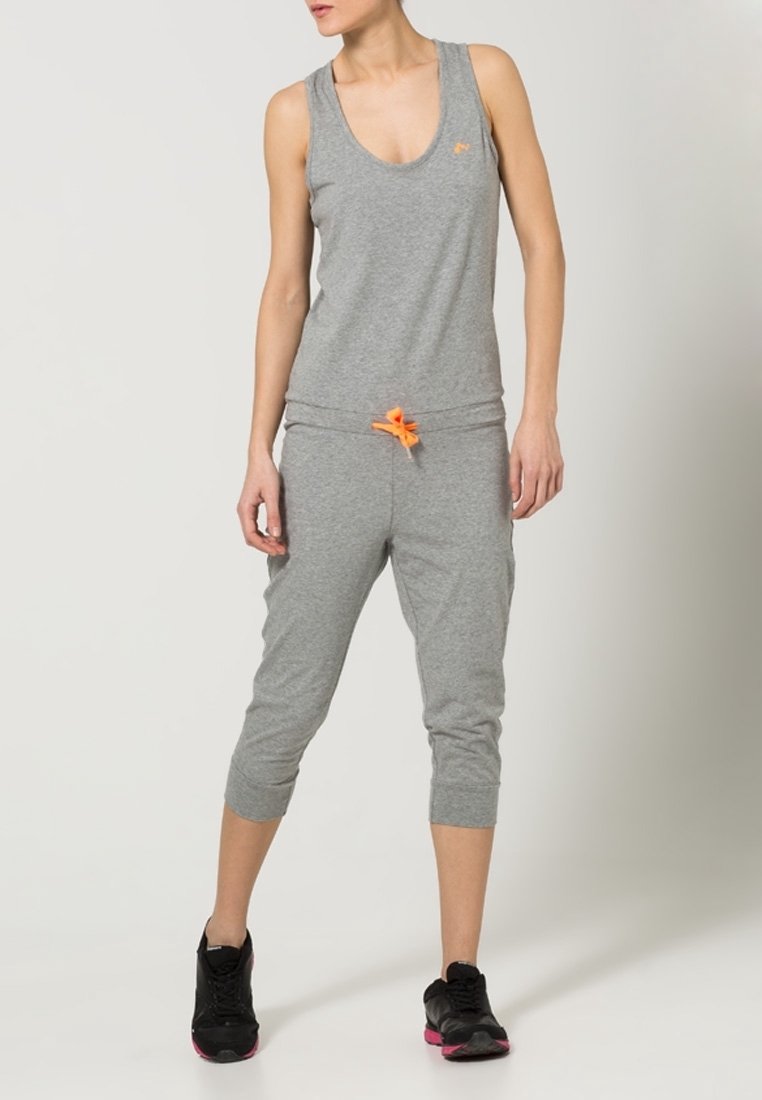 Femme portant un haut sans manches gris et un pantalon de survêtement court avec un cordon orange, des baskets noires avec des semelles roses, debout devant un fond uni.