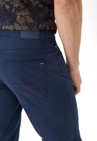 Marineblaue Hosen mit glatter Textur, einem hinteren Taschenfach mit Markenlogo und einem sichtbaren Taillentag, der das Design hervorhebt.