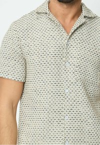 Chemise à manches courtes à boutons, de couleur beige clair, avec un motif texturé de petites boucles noires et crème. Elle possède un col et des boutons clairs.