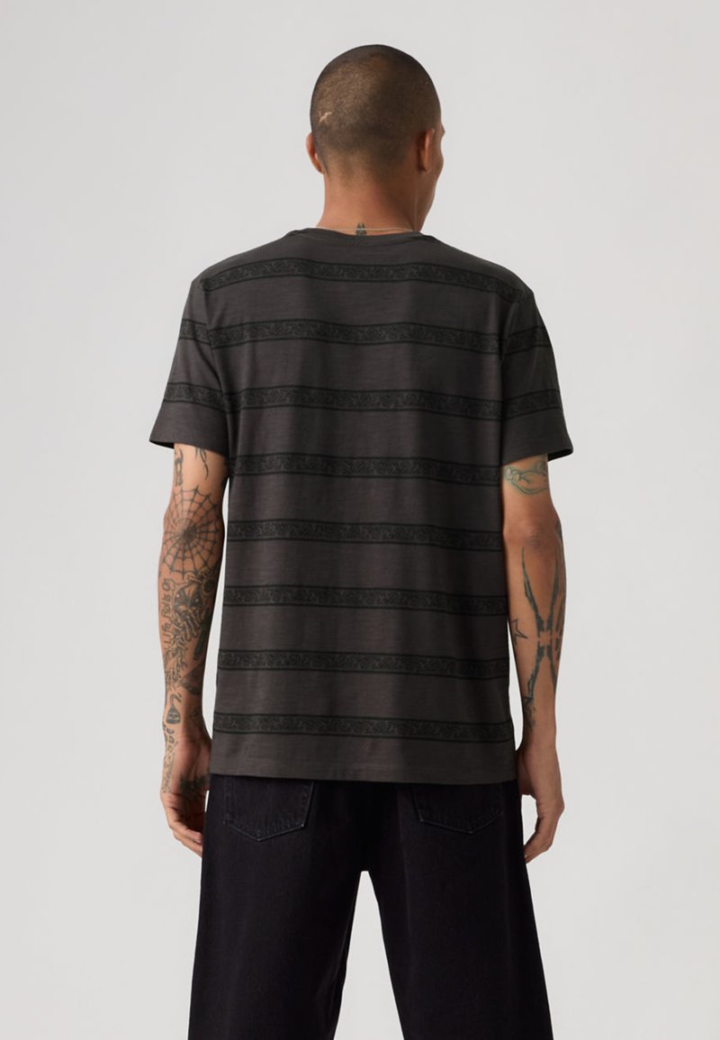 Homme avec des tatouages sur les bras et le cou, portant un t-shirt noir à manches courtes avec de subtiles rayures horizontales, vu de dos sur un fond uni.