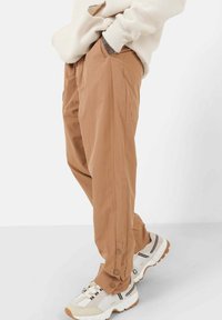 Lichtbruine, loszittende broek met knoopsluiting aan de zijkanten, gemaakt van een gladde stof. Gecombineerd met witte en beige sneakers.