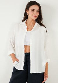 White, button-up shirt with a textured pattern; features a collar, rolled sleeves, and a relaxed fit. Paired with black drawstring pants.