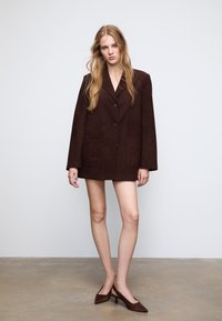Blazer oversize in suede marrone con due tasche frontali, chiusura a un bottone, revers larghi e maniche lunghe. Abbinato a scarpe col tacco alte di colore marrone.
