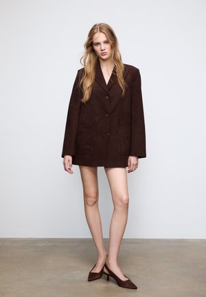 Brown suede oversized blazer with two front pockets, single button closure, wide lapels, and long sleeves. Paired with brown high-heeled shoes.