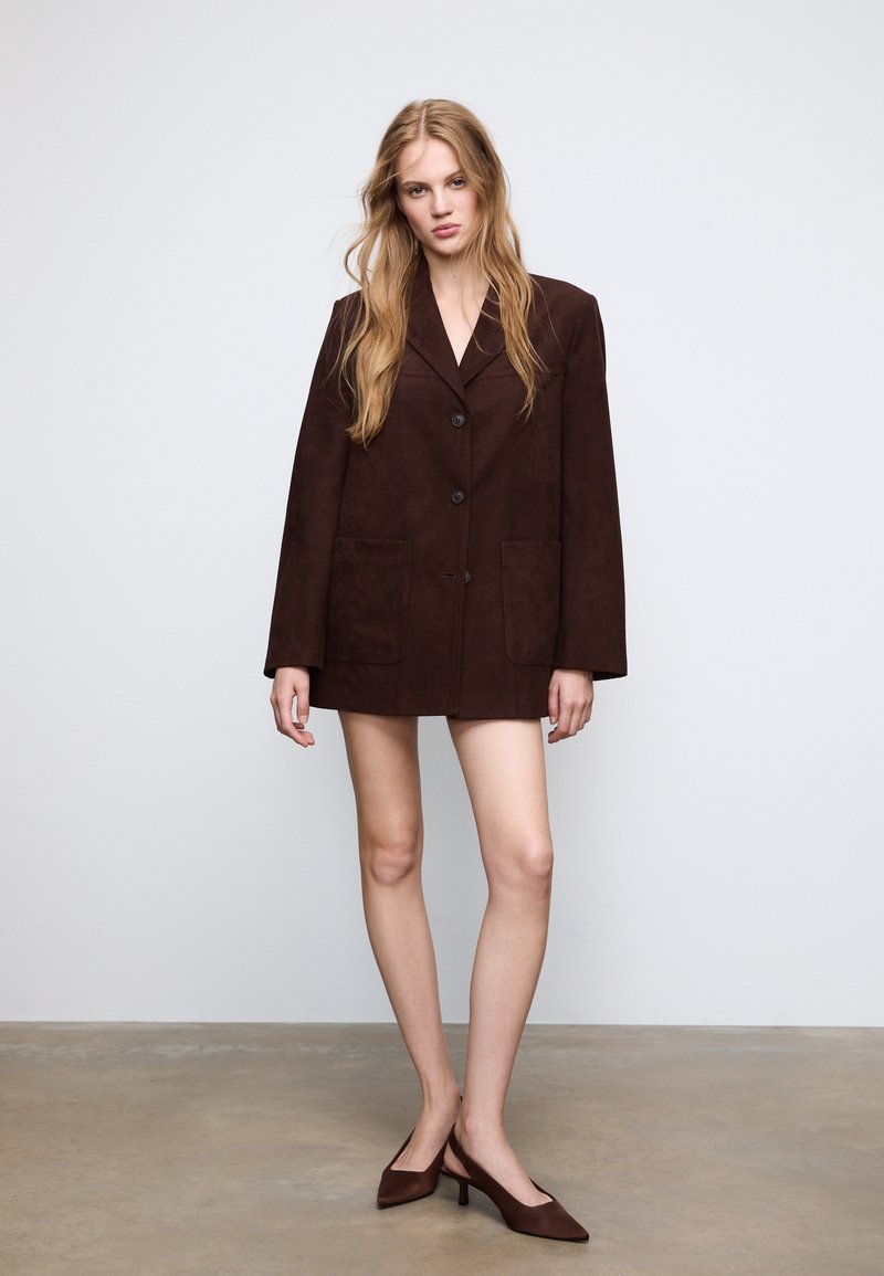 Blazer oversize in suede marrone con due tasche frontali, chiusura a un bottone, revers larghi e maniche lunghe. Abbinato a scarpe col tacco alte di colore marrone.