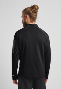 Schwarzes Langarmshirt aus strukturiertem Stoff, mit steigendem Kragen und grauen Akzentpaneelen an den Schultern. Enganliegend und leicht.
