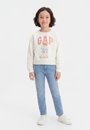 Παιδί με κοντά σκούρα μαλλιά που φοράει λευκό φούτερ με αρκουδάκι και το λογότυπο «GAP», ανοιχτό μπλε τζιν και λευκά αθλητικά παπούτσια, στέκεται όρθιο.