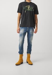 Svart t-shirt med en färgglad grafik av en tiger och ryttare, parat med slitna blå jeans och gula mocka ankelboots.
