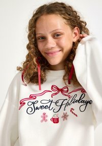 Hvid sweatshirt med teksten "Sweet Holidays" i sort kursiv, dekoreret med røde bånd og broderede jule-tema accenter.