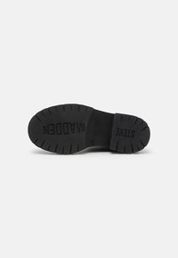 Suela de goma negra con textura, que presenta "STEVE MADDEN" grabado en letras mayúsculas. Forma redondeada con surcos pronunciados para un mejor agarre.