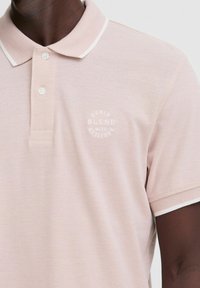 Lyserød polo t-shirt lavet af tekstureret stof, der har en hvid krave og manchetter, med et cirkulært logo, der står "DENIM BLEND WITH ATTITUDE."