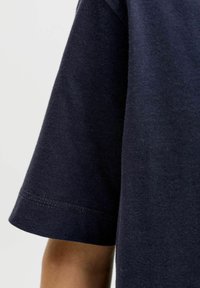 Κοντό μανίκι μπλε βαμβακερού t-shirt που φοριέται στο χέρ ι ενός ατόμου πάνω σε απλό ανοιχτό φόντο.