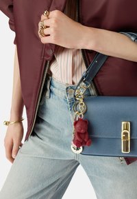 Sac à bandoulière en cuir texturé bleu avec un fermoir doré, agrémenté d'un charme ours rouge et de quincaillerie dorée. Porté avec un jean et une veste bordeaux.