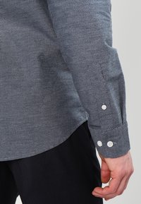 Košile s dlouhým rukávem v námořnické modři s texturovaným herringbone vzorem. Obsahuje bílé knoflíky na manžetách. Přiléhavý střih a zahnutý spodní lem.