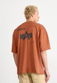 Bronzová bavlnená tričko s krátkymi rukávmi, s čiernym grafickým dizajnom a logom na chrbte. Jednoduchý a voľný strih.