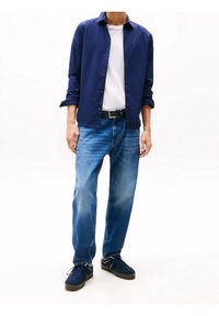 Camicia button-up blu navy, T-shirt bianca, jeans in denim azzurro chiaro, cintura nera, scarpe da tennis blu navy con accenti bianchi. Posizione eretta, sfondo neutro.
