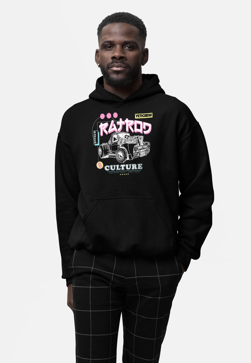 Man wearing black hoodie with vintage car and text "Ratrod Culture" graphic, paired with black pants featuring white grid lines.