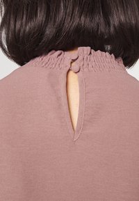 Blusa rosa con escote fruncido, espalda con abertura en forma de llave y cierre de botón, y textura suave y mate.