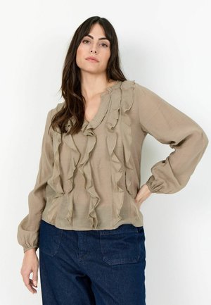 Mulher de cabelos longos e escuros, vestindo uma blusa bege com babados e calças azul-escuras, em pé com uma mão na anca contra um fundo branco.