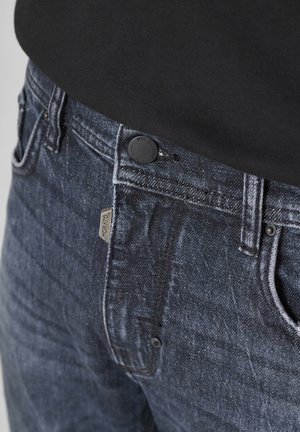 Tamno plave traperice imaju glatku teksturu, vidljivo šivanje, metalno dugme za zatvaranje i malu oznaku branda blizu džepa.