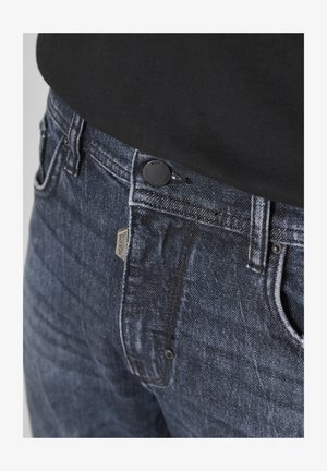 Un jean en denim bleu foncé présente une texture lisse, des coutures visibles, un système de fermeture à bouton en métal et une petite étiquette de marque près de la poche.