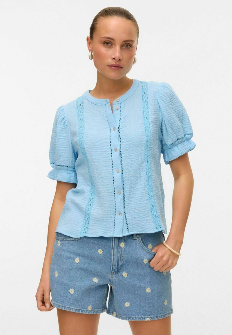 Blusa button-up azzurro chiaro realizzata in tessuto testurizzato, con dettagli in pizzo, maniche a sbuffo e colletto tondo, abbinata a pantaloni corti in denim ricamati.