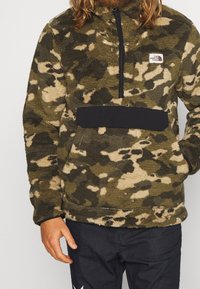 Sudadera polar camuflaje en verde y beige con cuello con cremallera, cinturilla negra y bolsillo. Textura suave con patrón distintivo.