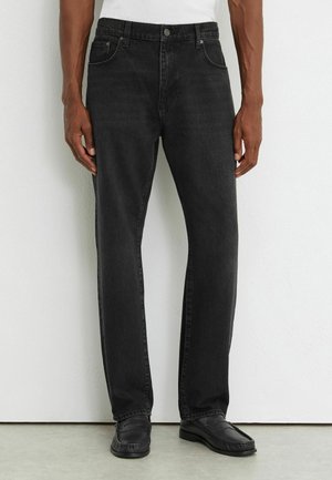 Homme portant un jean noir à jambes droites, des mocassins noirs et une chemise blanche, debout devant un mur clair uni.