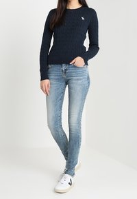 Kvinna klädd i en marinblå kabelstickad tröja med liten logotyp, ljusblå tajta jeans och vita sneakers med svarta detaljer, stående med en hand i fickan.