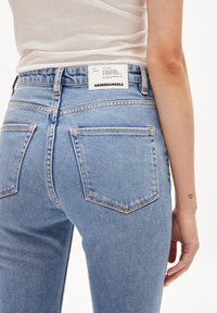 Vaaleat denim-farkut, joissa on korkea vyötärö. Takana on valkoinen brändilogo, ja perinteinen viisi-taskuinen muotoilu näkyvine ompeleineen.