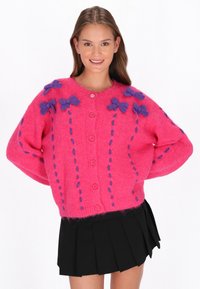 Cardigan roz tricotat, cu accente de fundă mov și detalii cusute, având un închidere cu nasturi și un croi larg, supradimensionat.