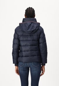 Giacca piumino navy con cappuccio, caratterizzata da sezioni a trapunta orizzontale, zip frontale e dettaglio del colletto con marchio. Abbinata a jeans blu.