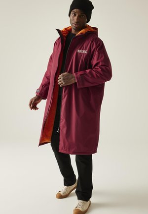 Mężczyzna w długiej bordowej kurtce Regatta z pomarańczowym polarem w środku, czarnych spodniach, białych trampkach i czarnej czapce, pozujący na jednolitym tle.