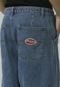 Denim pants, medium blue, featuring a single back pocket with a red and white branded patch. Classic straight-leg design with visible stitching.