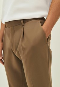 Pantaloni marroni con pieghe, caratterizzati da una superficie di tessuto liscia e tasche laterali. La cintura è dotata di passanti per cintura e presenta un design su misura.