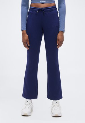 Person iført marineblå Adidas sweatpants med snøre i taljen og hvide Adidas sneakers, stående med armene afslappet ned langs siden.