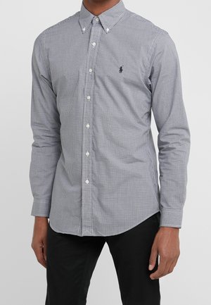 Homme portant une chemise slim à manches longues avec des boutons, à petit motif à carreaux noir et blanc, et un pantalon noir.