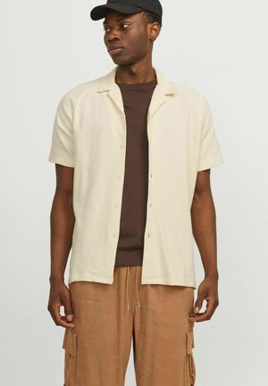 Kortærmet, lysebeige skjorte med knaplukning og krave, båret over en brun t-shirt; ledsaget af brune cargo-shorts.