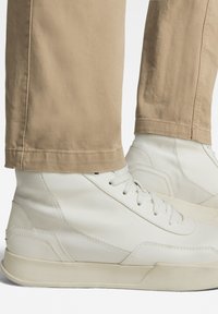 White high-top sneakers made of leather, featuring textured panels and flat laces, paired with light beige pants cropped at the ankle.