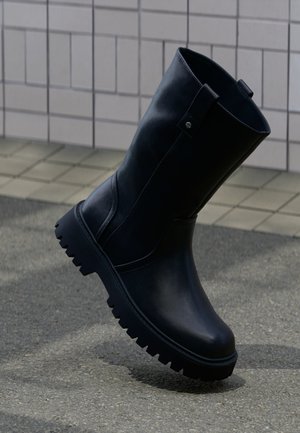 Bottes en cuir noires mi-mollet avec semelle crantée épaisse devant un mur carrelé et un trottoir.