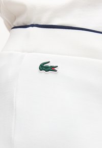 Λευκή ζώνη ενδύματος με ναυτικό μπλε ρίγα και πράσινο λογότυπο κροκόδειλου ραμμένο κοντά στο κέντρο σε λείο ύφασμα.