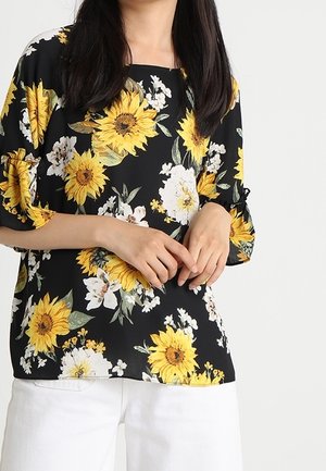 Femme portant un chemisier noir avec un imprimé tournesol jaune vif et fleurs blanches, associé à un pantalon blanc, mains doucement jointes.