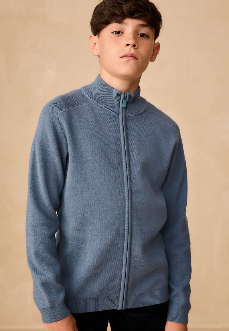 Ragazzo con capelli scuri corti indossa un maglione blu con zip, in piedi davanti a uno sfondo beige semplice, guarda dritto in avanti con espressione neutra.