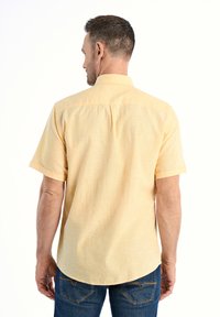 Korte mouwen gele shirt met een gestructureerde stof, knoopsluiting kraag en afgeronde zoom. Draagt het met blauwe jeans, gezien van achteren.