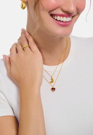 Goldfarbene, geschichtete Halsketten mit einem herzförmigen Anhänger mit rotem Edelstein und einem Mond-Anhänger. Getragen zu einem schlichten weißen T-Shirt.