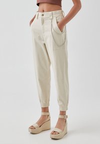 Crème-kleurige katoenen broek met een elastische tailleband, zijzakken en een zilveren kettingdetail. Gecombineerd met beige instap sandalen.