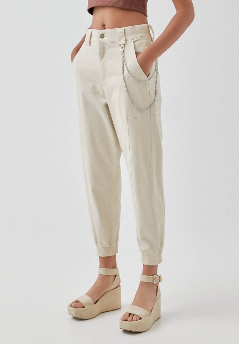 Crème-kleurige katoenen broek met een elastische tailleband, zijzakken en een zilveren kettingdetail. Gecombineerd met beige instap sandalen.