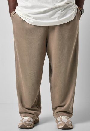Persona vistiendo pantalones beige holgados, zapatillas blancas y una camiseta blanca con las manos en los bolsillos, de pie sobre un suelo gris.